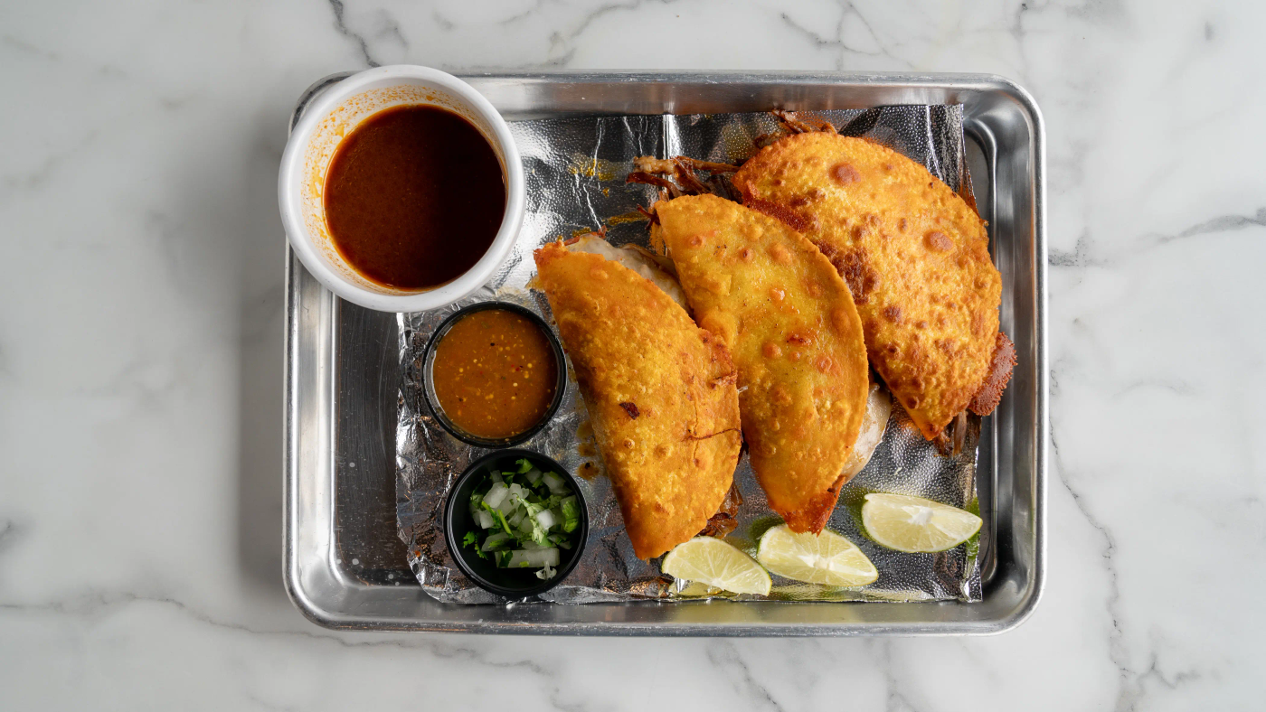 Three soft corn tortillas filled with birria-style cooked beef. Served with a side of broth (consomé) for dipping, cilantro, onions, lime, and homemade spicy sauce on the side